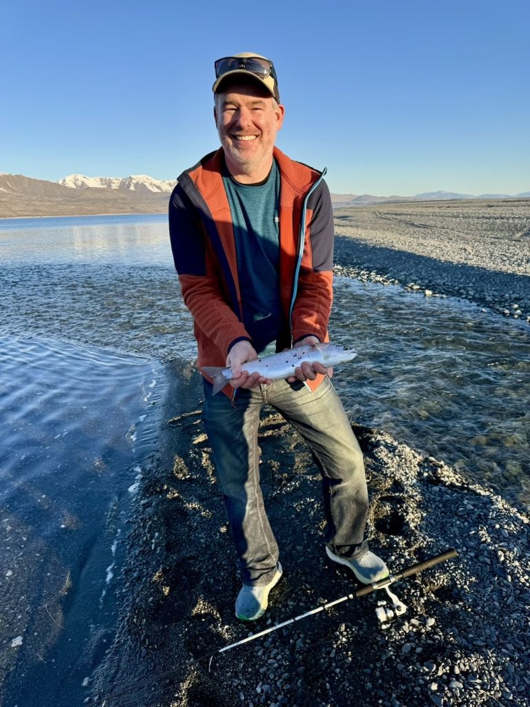 Smiling man holding a fish he caught in a river with mountains in the background.