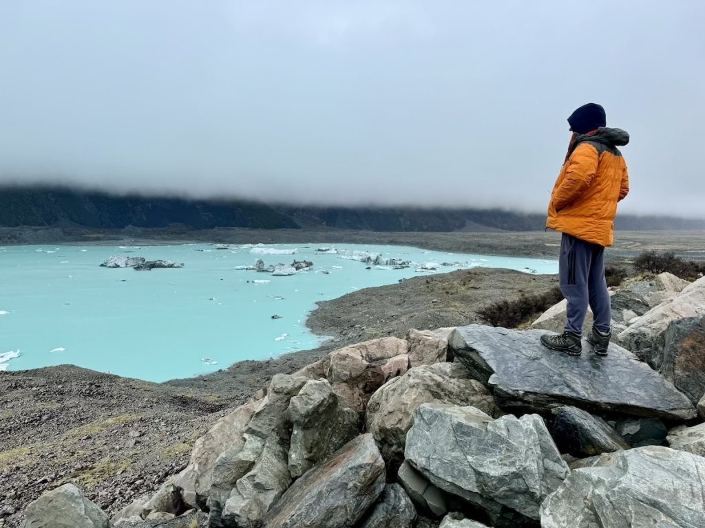 Person in orange jacket looks at glacial lake with icebergs on cloudy day.