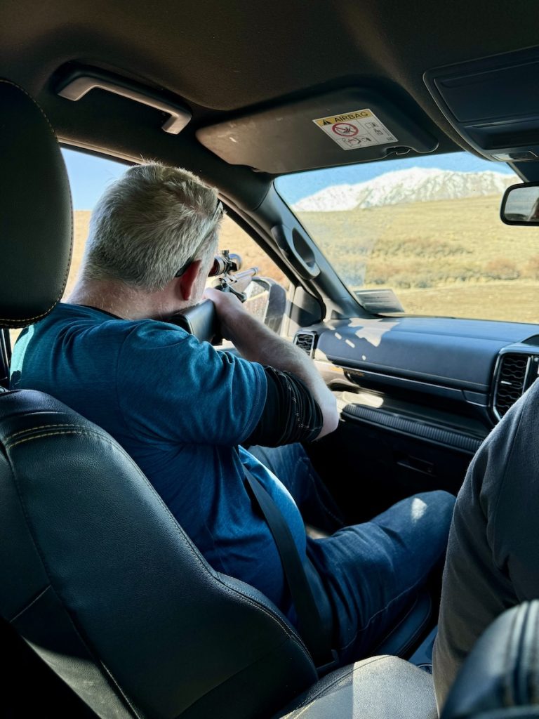 Man aims rifle out car window at distant landscape with mountains.