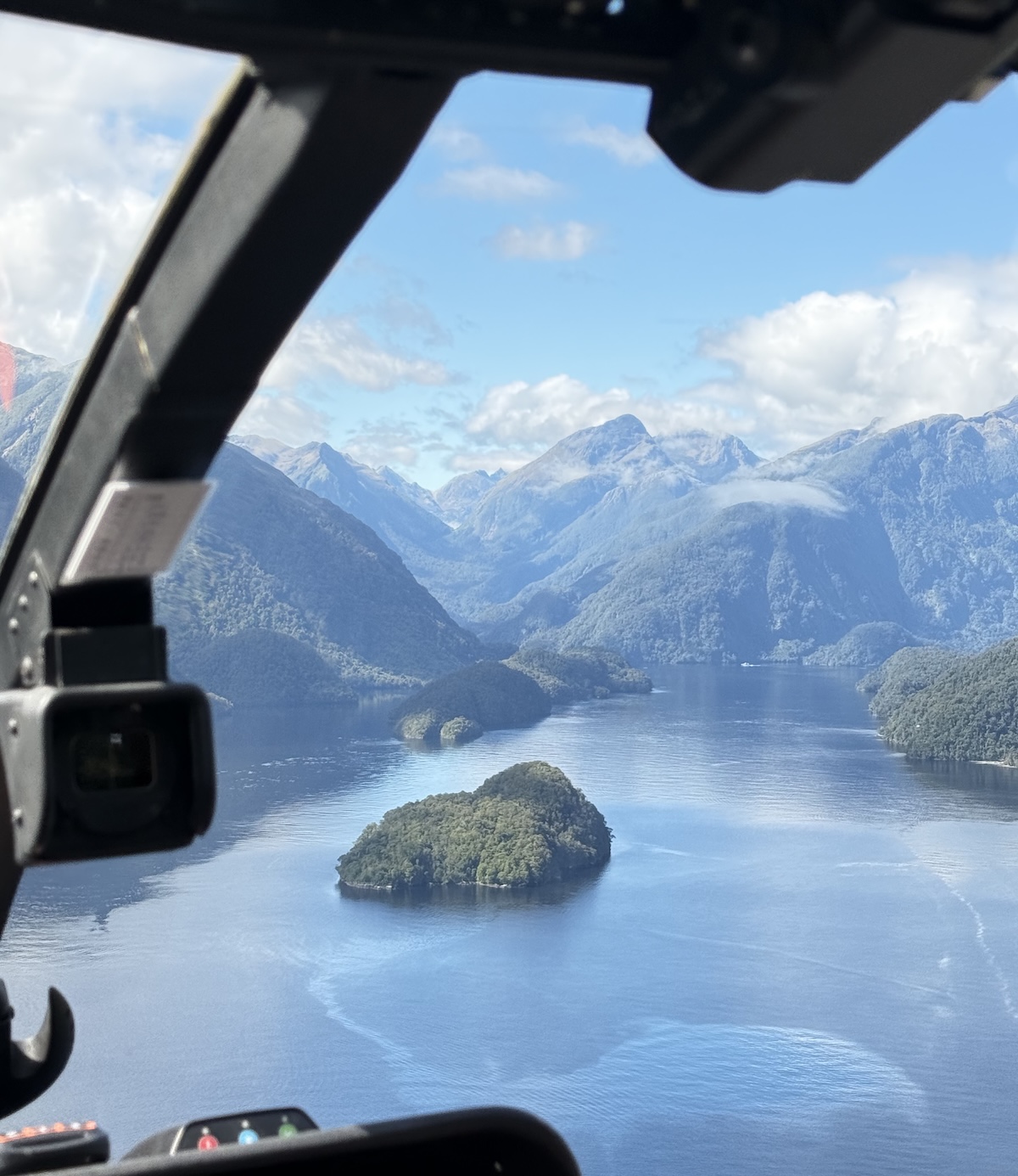 A view from a helicopter cockpit looking out over a calm body of water surrounded by forested mountains and small islands.
