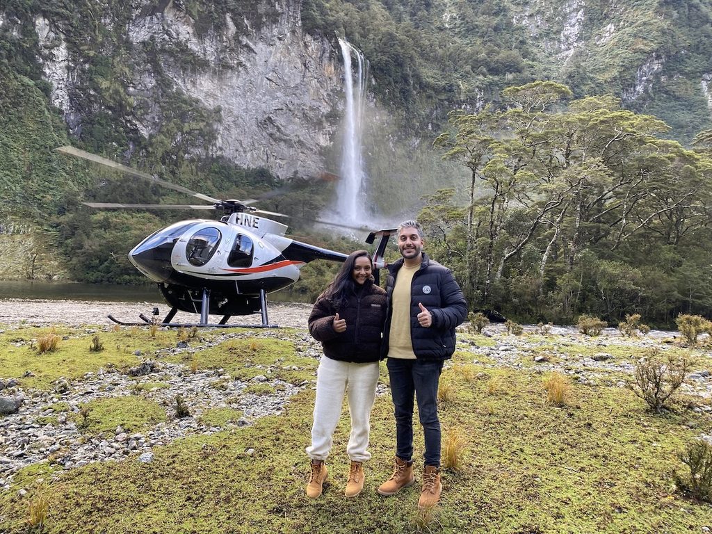 A man and a woman stand giving thumbs up in front of a helicopter parked on rocky ground near a waterfall and forest.