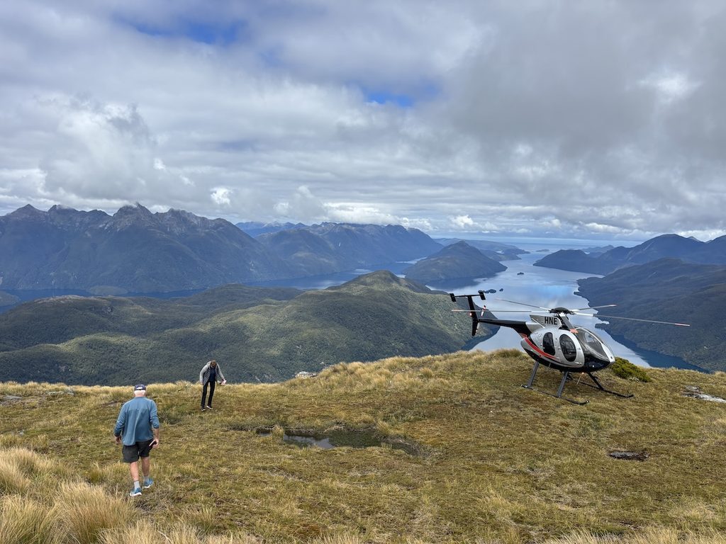 Two people walk on a grassy mountain ridge near a parked helicopter overlooking a large lake and distant mountains.