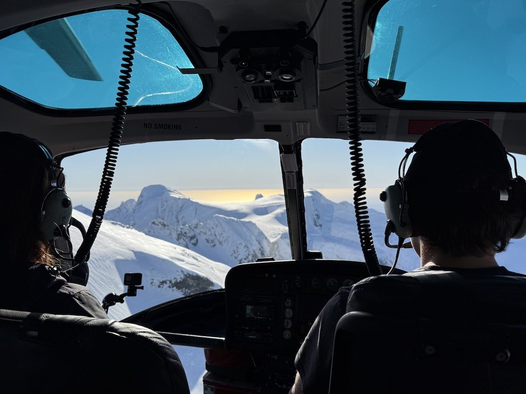 Two people wearing headsets sit in the cockpit of a helicopter looking out at snow-covered mountains.