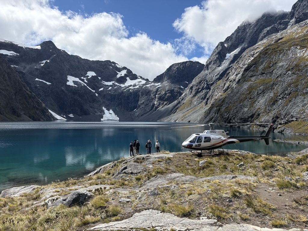 A helicopter is parked on a rocky shore beside a blue mountain lake with a group of people standing nearby.