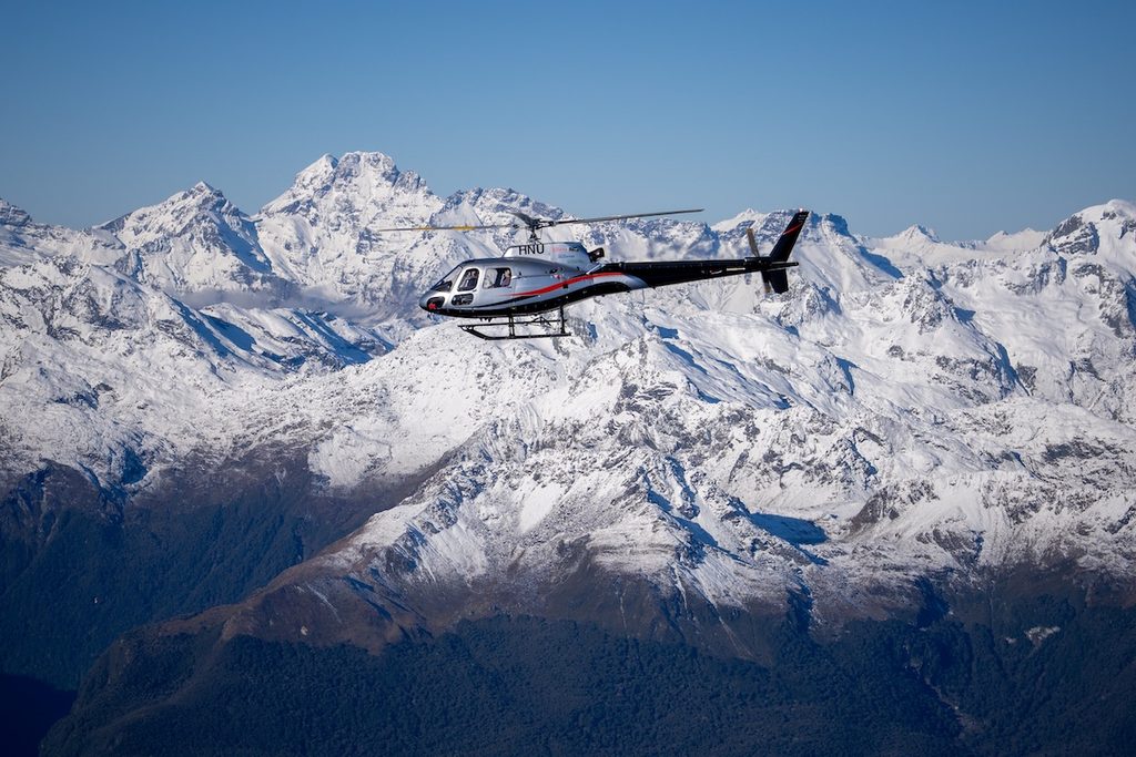 A silver and black helicopter flies in front of snow-covered mountain peaks under a clear blue sky.